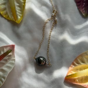 May include: A gold chain bracelet with a large, dark gray pearl pendant. The bracelet is on a white surface, surrounded by yellow and pink flowers. The bracelet has a small clasp and a few small white pearls.