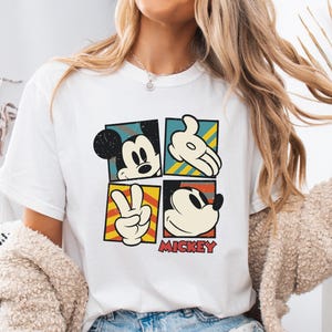 May include: White t-shirt with a vintage-style graphic of Mickey Mouse in four different poses. The graphic is in a colorful block design with yellow, red, blue, and green accents. The text "MICKEY" is printed below the graphic.
