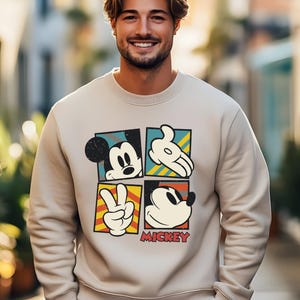 May include: A beige sweatshirt with a vintage-style graphic of Mickey Mouse in four different poses. The graphic is in a colorful block design with the word "MICKEY" printed below.