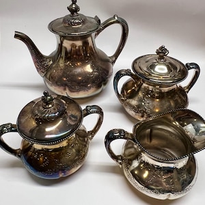 May include: A vintage silver tea set, including a teapot, sugar bowl, creamer, and a small covered bowl. The set features ornate floral detailing and curved handles. The teapot has a spout and a lid with a decorative finial.