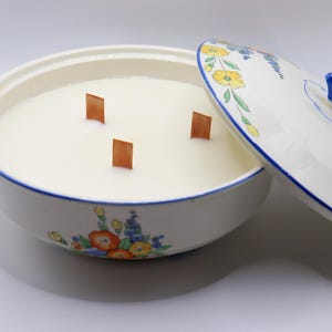 May include: A large, round candle in a decorative ceramic bowl with a lid. The bowl is white with blue trim and floral designs. Three wooden wicks are visible in the creamy wax. The lid is partially open, revealing the candle.