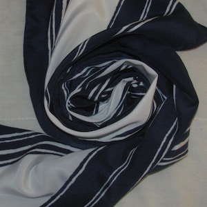 Peut inclure: Un foulard bleu marine et blanc, plié en spirale. Le foulard présente un motif répétitif de rayures blanches sur un fond bleu marine. Le foulard est fait d'une matière soyeuse.