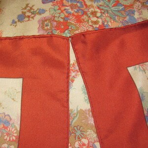 May include: A square scarf featuring a cream-colored background with a multi-colored floral pattern in shades of red, blue, and green. The scarf has a wide, solid rust-red border along the edges with visible stitching along the hemline.