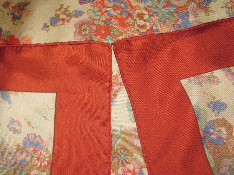 May include: A square scarf featuring a cream-colored background with a multi-colored floral pattern in shades of red, blue, and green. The scarf has a wide, solid rust-red border along the edges with visible stitching along the hemline.
