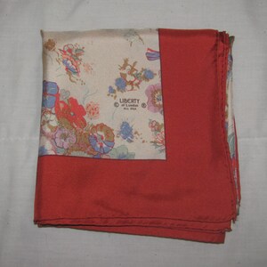 May include: A square silk scarf featuring a cream-colored section with a multi-colored floral pattern and a solid red border. The text LIBERTY of London ALL SILK is printed on the cream section. The scarf is folded to show both patterns.