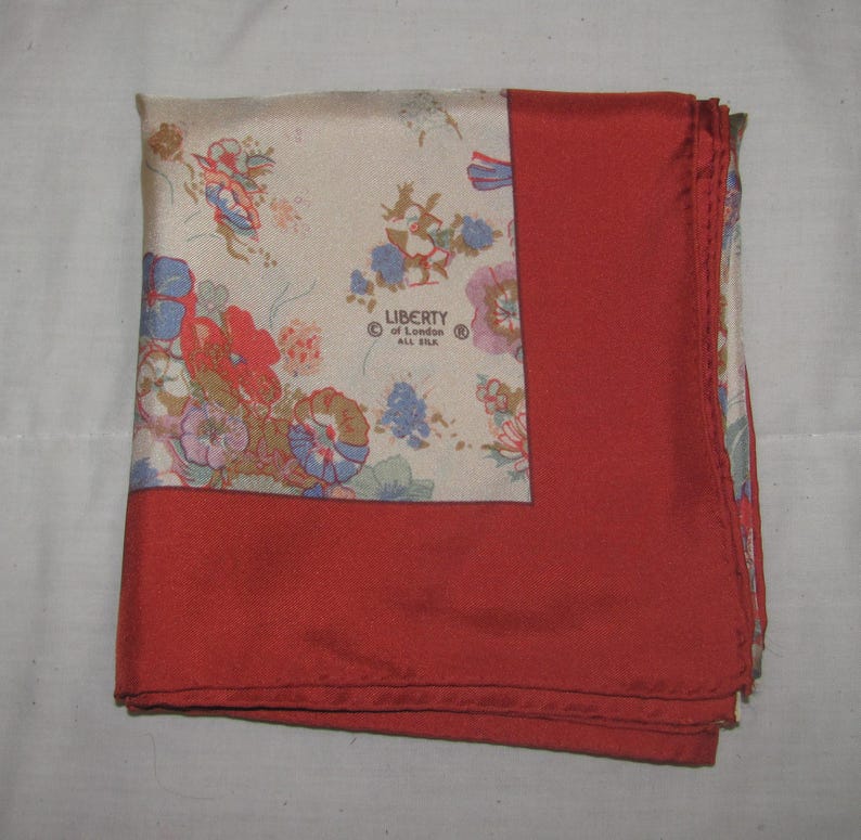 May include: A square silk scarf featuring a cream-colored section with a multi-colored floral pattern and a solid red border. The text LIBERTY of London ALL SILK is printed on the cream section. The scarf is folded to show both patterns.