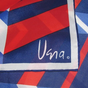 May include: A silk scarf with a geometric pattern in red, white, and blue. The design features angled stripes and shapes, with the word "Una" in white script in the corner. The scarf has a shiny finish.