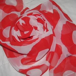 Peut inclure: Une écharpe à motifs rouge et blanc avec un fond blanc et des pois rouges. L'écharpe est en tissu transparent et est pliée de manière décorative. Le nom de la marque "Viga" est imprimé en rouge sur l'écharpe.
