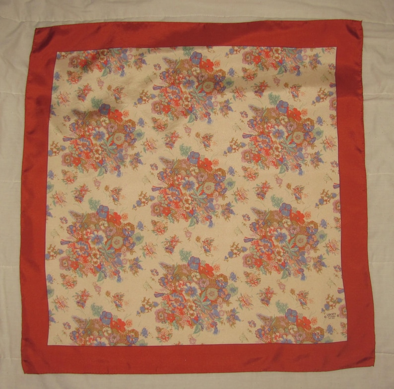 May include: A square scarf featuring a cream-colored center with a repeating floral pattern in shades of red, blue, and green. The scarf is framed by a solid, wide red border along all four edges, creating a distinct contrast with the central print.