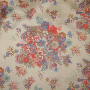 May include: A beige fabric featuring a repeating floral pattern with clusters of flowers in shades of red, blue, purple, and muted green. The design consists of various small blossoms and leafy sprigs scattered across the light tan background.