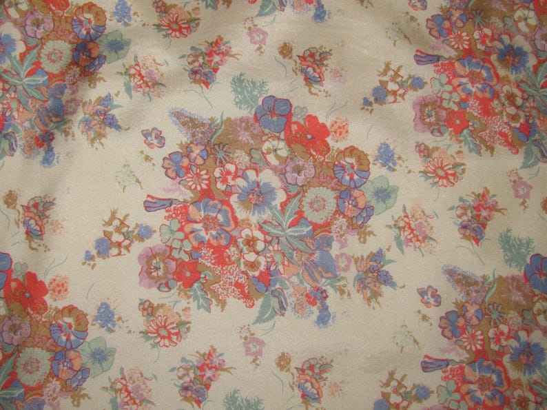 May include: A beige fabric featuring a repeating floral pattern with clusters of flowers in shades of red, blue, purple, and muted green. The design consists of various small blossoms and leafy sprigs scattered across the light tan background.