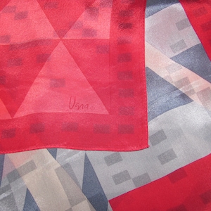 May include: A red and geometric patterned scarf with a red border. The scarf features a design of triangles and squares in shades of red, grey, and white. The signature "Usna" is visible in the corner.