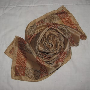 May include: A folded silk scarf in shades of tan, brown, and rust. The scarf has a repeating pattern of small dots in rust and grey, arranged in vertical stripes along the edges. The scarf is arranged in a spiral, showcasing its texture and design.