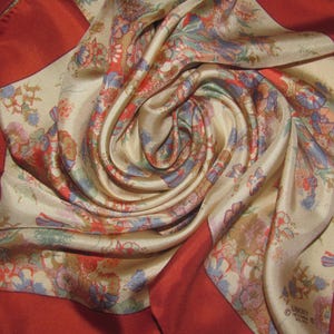 May include: A square silk scarf featuring a cream-colored center with a multi-colored floral pattern in shades of blue, red, and green. The scarf is framed by a wide, solid deep red border and includes the printed text Liberty of London on one corner.