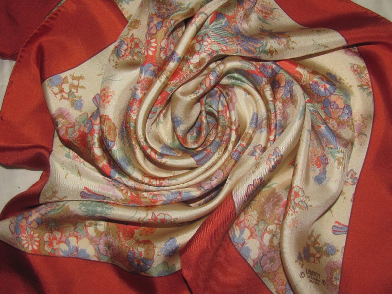 May include: A square silk scarf featuring a cream-colored center with a multi-colored floral pattern in shades of blue, red, and green. The scarf is framed by a wide, solid deep red border and includes the printed text Liberty of London on one corner.