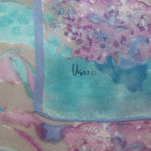 May include: A close-up of a silk scarf with a watercolor design. The scarf features a blend of teal, lavender, and gray hues with floral and abstract patterns. The artist's signature, "Usna", is visible in black.