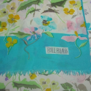 May include: A floral patterned scarf with a turquoise border and fringe. The scarf features a variety of colorful flowers in yellow, pink, and blue against a white background. The brand name "BILL BLASS" is printed on the turquoise border.