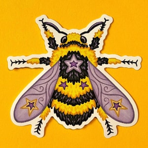 May include: A die-cut sticker featuring a stylised bumblebee with a fuzzy yellow and black striped body. The bee has light purple wings decorated with gold-outlined purple stars and a large purple star on its back. It has black legs and antennae.