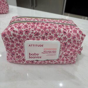 Peut inclure: Une pochette en tissu rectangulaire avec un motif floral rose et blanc et une fermeture éclair sur le dessus. Au centre se trouve un couvercle distributeur en plastique blanc marqué ATTITUDE baby leaves lingettes 99% d'eau.