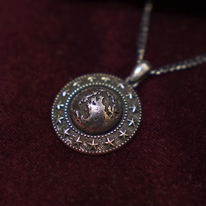 May include: Silver pendant necklace with a round pendant featuring a detailed world map design. The pendant is encircled by a ring of stars. The necklace is displayed on a dark red velvet surface, with a partially visible gift box.