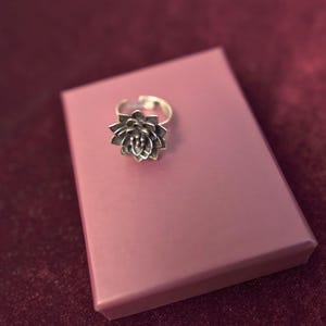 Puede incluir: Un anillo de plata con un dise&ntilde;o de flor. El anillo est&aacute; en una caja rosa.