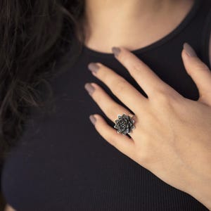 Puede incluir: Un anillo de plata con un dise&ntilde;o de flor negra. El anillo est&aacute; en el dedo de una persona.