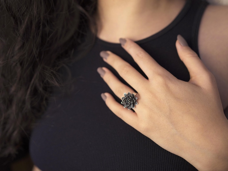Puede incluir: Un anillo de plata con un dise&ntilde;o de flor negra. El anillo est&aacute; en el dedo de una persona.