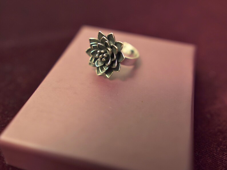 Puede incluir: Un anillo de plata con un dise&ntilde;o floral detallado. El anillo est&aacute; sobre una superficie rosa.