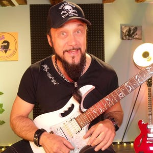 May include: A man wearing a black t-shirt and a black baseball cap with a skull and crossbones logo, holding a white electric guitar with a detailed fretboard. A red electric guitar is in the background.