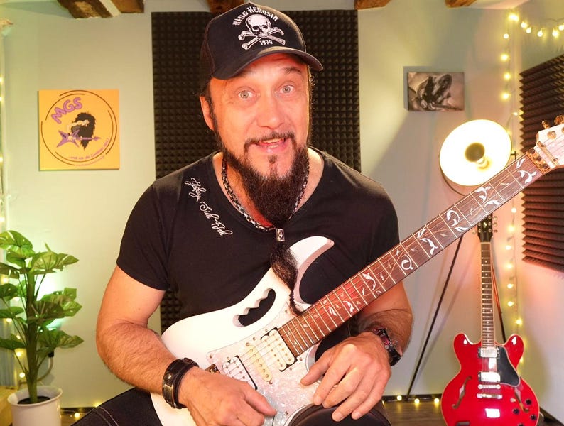 May include: A man wearing a black t-shirt and a black baseball cap with a skull and crossbones logo, holding a white electric guitar with a detailed fretboard. A red electric guitar is in the background.