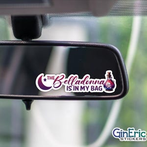 May include: A sticker with the text "The Belladonna is in my bag" in a cursive font, with a purple crescent moon and star design. A cartoon potion bottle with a skull is also featured. The sticker is placed on a car's rearview mirror.