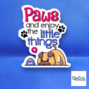 May include: A die-cut sticker featuring a tan cartoon dog lying down next to a blue food bowl. The text reads Paws and enjoy the little things in pink and blue lettering, accented by two black paw prints and a small pink speech bubble with a white heart.