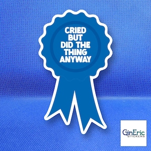 May include: A blue award ribbon sticker with a white border and white text that reads CRIED BUT DID THE THING ANYWAY. The sticker features a circular top with a scalloped edge and two downward-pointing ribbon tails at the bottom center.