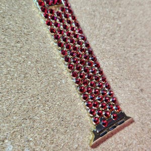 以下が含まれることがあります： 赤いクリスタルデザインの金色の腕時計バンド。バンドは金属製で、留め金で留まります。