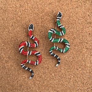 May include: A pair of snake-shaped earrings with rhinestones. One earring is red, black, and white, and the other is green, black, and white.