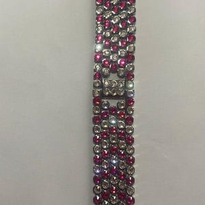 May include: A dazzling watch strap with a silver-tone metal base, embellished with alternating rows of pink and clear crystals. The strap has a rectangular crystal feature in the centre and is designed for a smartwatch.
