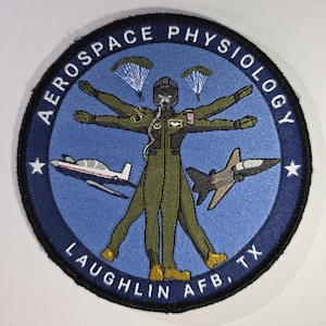 May include: A circular patch with the words "AEROSPACE PHYSIOLOGY" and "LAUGHLIN AFB, TX" in white text on a blue background. The patch features a pilot in a flight suit with outstretched arms, two parachutes, two airplanes, and two stars.