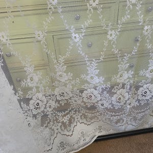 May include: White lace curtain panel with a floral pattern. The lace is delicate and has a scalloped edge.