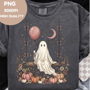 May include: A charcoal gray t-shirt featuring a graphic of a white ghost sitting on a wooden swing. The design includes a pink balloon, a crescent moon, bats, stars, and a collection of pink and orange pumpkins surrounded by various flowers.