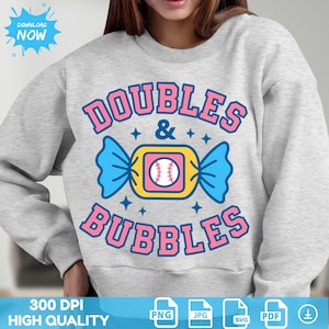 May include: A pale grey sweatshirt featuring the text "DOUBLES & BUBBLES" in pink. The design includes a baseball within a candy wrapper, with blue and yellow details. The sweatshirt also has a "DOWNLOAD NOW" graphic.