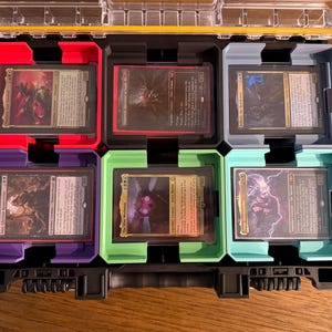 May include: A black and yellow plastic case with six compartments, each holding a trading card. The compartments are coloured red, purple, green, blue, and grey. The cards have various illustrations and text.