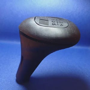 Op de afbeelding: Een zwarte versnellingspookknop met de cijfers 1, 2, 3, 4 en 5 zichtbaar. De bovenkant van de knop heeft een rechthoekige inkeping met een reeks lijnen. De knop staat op een blauwe achtergrond.