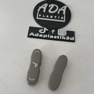 May include: Two gray, oblong-shaped plastic objects with rounded ends and a textured surface. One has a curved line and a small arrow. The image also shows a black circular logo with the text "ADA PLASTIK" and a black rectangular logo with social media icons and the text "Adaplastik3d".