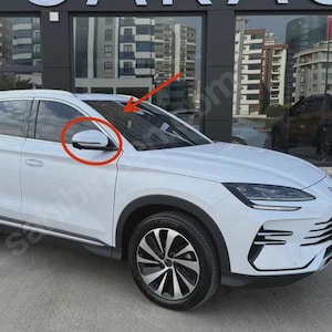 May include: A white SUV with black accents, featuring a side mirror and a black and silver wheel. The vehicle is parked in front of a building with large windows, reflecting the surrounding cityscape.