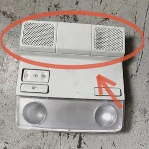 Puede incluir: Una consola superior interior de coche blanca con dos rejillas de altavoz, dos luces redondas y tres botones. La consola rectangular tiene una superficie texturizada.