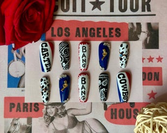 May include: A set of ten artificial nails with various designs. Some nails are white with black patterns, others are blue with gold accents, and some have text like "CARTER", "BANG", and "COWBOY". The nails are arranged on a pink background.