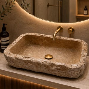 Puede incluir: Un lavabo rectangular tallado en piedra beige natural con un exterior rugoso y texturizado y una cubeta interior lisa. Cuenta con un grifo de oro cepillado y un tapón de drenaje circular a juego, colocado sobre una encimera de piedra.