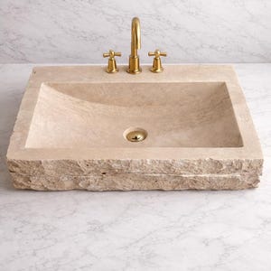 Op de afbeelding: Een rechthoekige, beige stenen wastafel met een ruwe rand. De wastafel heeft een gebogen bekken en een goudkleurige kraan en armaturen. De wastafel staat tegen een witte marmeren achtergrond.
