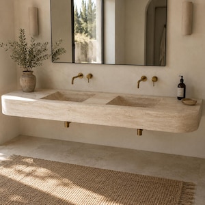 May include: A wall-mounted double vanity sink crafted from beige travertine stone with a natural textured finish. It features two integrated rectangular basins, two sets of brushed gold wall-mounted faucets, and matching gold plumbing fixtures underneath.