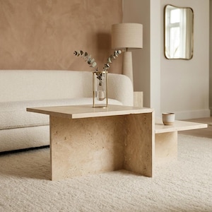 May include: A two-tiered coffee table constructed from beige travertine stone with a porous, natural texture. The table features a taller rectangular section and a lower, wider section, both supported by solid slab legs. A glass vase sits on top.
